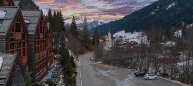 Dolomites, İtalya 'da bir dağ köyü, ahşap bir otel, kilise kulesi, karlı dağlar, orman yamaçları ve renkli bir günbatımı gökyüzü..