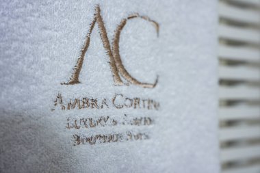 Üzerinde Ambra Cortina Lüks ve Fashion Boutique Hotel logosu olan altın işlemeli beyaz havlu. Arkaplanda görünen bulanık yüzey.