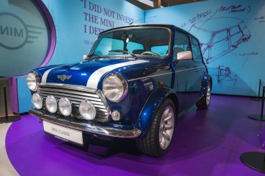 Klasik Mini Cooper. Beyaz yarış çizgileri ve rali ışıkları BMW Müzesi 'nde sergileniyor. Üzerinde mini bir plan olan bir duvara karşı dizilmiş..