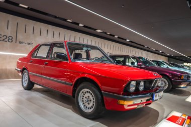 1981 yılından canlı kırmızı bir BMW E28 sedan BMW Müzesi 'ndeki parlak bir platformda, bir zaman çizelgesi ve diğer BMW modelleri arka planda görünür..