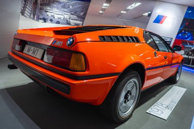 BMW Müzesi 'nde sergilenen parlak turuncu 1978 BMW M1 arka camlar, çift yuvarlak stop lambaları ve tarihsel gösterileri olan modern bir iç mekan..