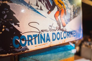 Karlı dağlara ve yemyeşil ağaçlara karşı dizilmiş turuncu pantolonlu bir snowboardcunun resimli posteri. Cortina Dolomitlerini destekleyen cesur metinler..