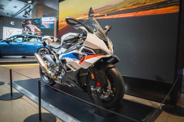 Platformda beyaz, mavi ve kırmızı bir BMW motosiklet, arka planda bir BMW araba ve gün batımında bir sürücüyü gösteren bir ekran..