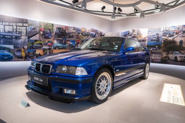 Münih 'teki BMW Müzesi' nde mavi bir 1992 BMW 3 serisi sergilendi. Arka planda BMW araç resimleri kolajlı eğri bir duvar var..