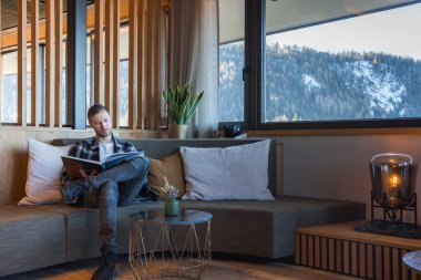 Bir adam, modern bir otel odasındaki minderli bir kanepede ahşap aksanlı, saksı bitkisi ve kar tozlu dağlara bakan büyük pencereli bir kitap okuyor..