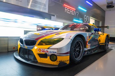 Münih 'teki BMW Müzesi' nde sarı, kırmızı ve mavi üniformalı bir BMW yarış arabası sergileniyor. Neon ışıklı duvar ve hazır set GO dinamik bir arkaplan ekler.