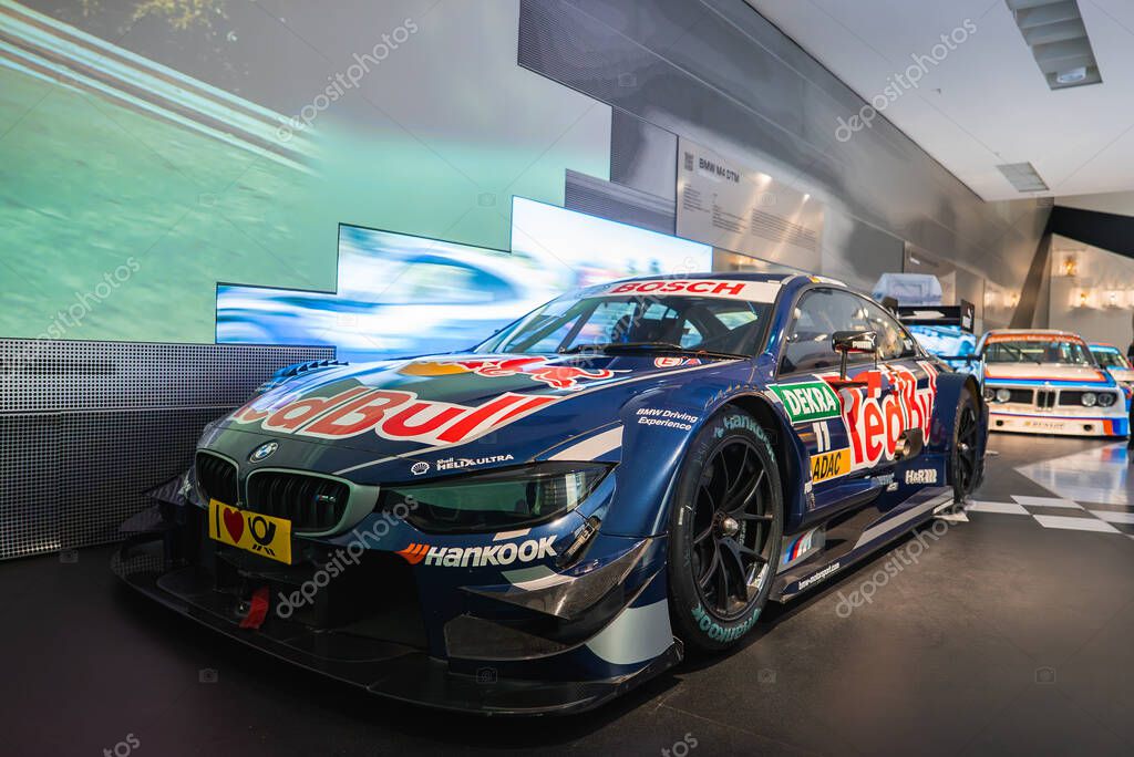 A BMW race car with Red Bull livery displayed in the BMW Museum, Munich. Sponsors logos and a racetrack themed wall enhance the motorsport exhibit.