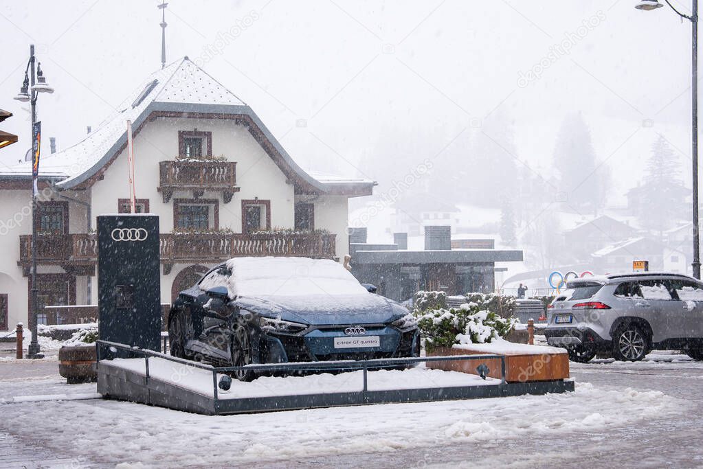 A modern Audi car on a snowy platform in Cortina dAmpezzo, Italy. An Alpine style building and faint Olympic rings appear in the wintery background.