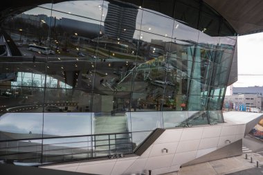 Münih 'teki BMW Welt binasında yakındaki binalar, ağaçlar ve araçları yansıtan geniş pencereli gösterişli ve açılı mimari yer alıyor..