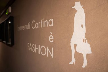 Bej desenli duvar Benvenuti Cortina ve FASHION yazıları ve şapka ve çanta taşıyan beyaz bir kadın silueti içeriyor..