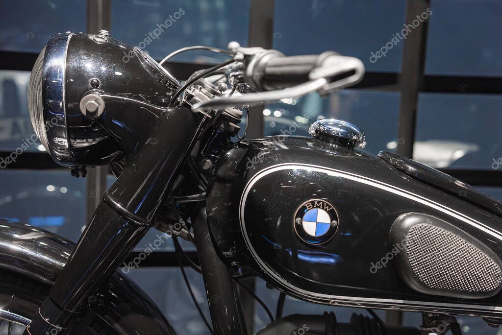 A vintage black BMW motorcycle with chrome accents and a rounded headlight, set against a modern glass paneled wall in an indoor exhibition space.