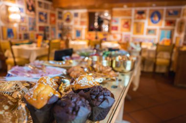 Sıcak aydınlatılmış bir yemek alanında, içinde hamur işleri ve ikramlar olan bir kahvaltı büfesi. Sarı döşemeli sandalyeler ve çerçeveli sanat eserleri butik otel alanını süslüyor..