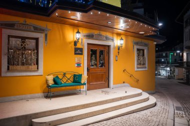 Cortina 'daki Ambra Oteli' nin girişinde sarı bir cephe, dekoratif detaylar, fenerler, yeşil bir bank, taş basamaklar ve geceleri kaldırım taşı bir cadde bulunur..