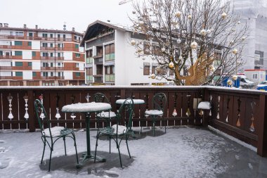 Açık teras, karla kaplı masa ve sandalyeler, ahşap korkuluklar, altın süslemeli şenlik ağacı, ve arka planda konut binaları..