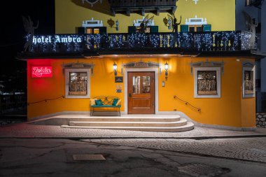Cortina 'daki Ambra Oteli' nin girişinde sarı bir cephe, melek heykelleri, tel lambaları, kırmızı neon bir tabela ve ön planda kaldırım taşları bulunur..
