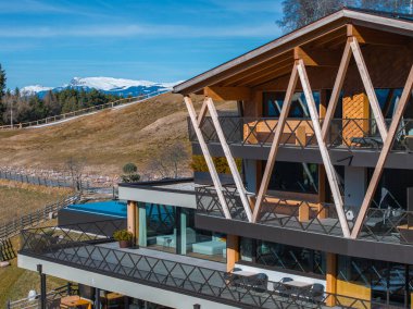 Çağdaş otel, üç köşeli ahşap kirişler, cam pencereler, teraslar, karlı dağlar, çimenli alanlar ve Güney Tyrol 'da açık mavi bir gökyüzü..
