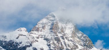 Alplerdeki Matterhorn dağında engebeli, karla kaplı yamaçlar, üçgen bir zirve ve ve ve bulutlar berrak mavi gökyüzüne karşı..