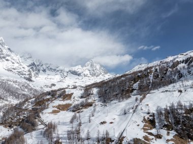 Matterhorn yakınlarındaki yamaçlarda kayak pistleri, dağınık ağaçlar, dolambaçlı bir yol ve Alpler 'de parçalı bulutlu bir gökyüzü vardı..