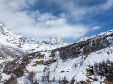 Karlı yamaçlar, dağınık ağaçlar ve Matterhorn Dağı 'nın yakınındaki dolambaçlı yollar açık mavi gökyüzünün altında dağılmış bulutlar Alplerde..