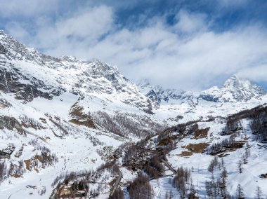 Kar yamaçları, dağınık ağaçlar ve dolambaçlı yollar, Cervino Kayak Cenneti yakınlarındaki Alpler 'de parçalı bulutlu bir gökyüzünün altında yükselen Matterhorn' u çevreledi..