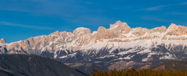 İtalya 'nın Güney Tyrol kentindeki Dolomite sıradağlarında açık mavi gökyüzü altında pürüzsüz kar örtülü zirveler, güneşli yamaçlar ve yoğun ormanlar bulunur..