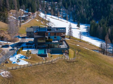 Camdan pencereleri, düz çatısı ve havuzu olan çağdaş bir otel. Güney Tyrol 'da bir yamaçta. Ormanlar, tepeler ve seyrek kar parçalarıyla çevrili.