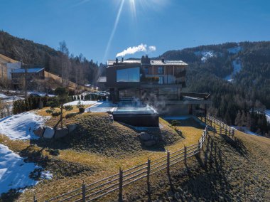 Camdan pencereleri ve düz çatısı olan çağdaş bir otel. Çimenli bir tepenin üzerinde, karlı dağlar ve ormanlarla çevrili dumanı tüten bir sonsuz havuz var..