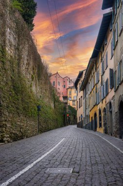 İtalya 'nın Bergamo şehrinde tarihi binalar, pastel cephelerle çevrili bir kaldırım taşı caddesi ve kullanım hatları olan sıcak bir günbatımı gökyüzünün altındaki pencereler..