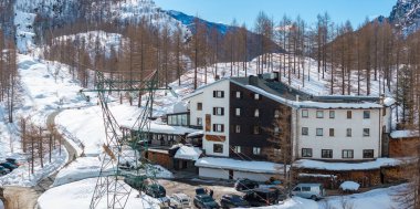 Matterhorn yakınlarında beyaz ve koyu renk ahşap dış görünüşlü, karla kaplı tepeler, kayak asansörü, park edilmiş arabalar ve engebeli tepelerle çevrili rahat bir otel..