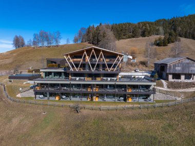 Güney Tyrol, İtalya 'da modern bir otel, ahşap üçgen dizayn, büyük cam pencereler, teraslar, çimenli tepeler, seyrek ağaçlar ve kırsal bir ahır..