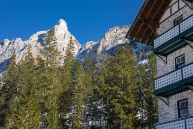 Engebeli Dolomite tepeleri, kar tozu dolu Evergreen 'lerin çerçevelediği açık mavi gökyüzüne karşı yükselir. Alp tarzı bir bina olay yerine kırsal ayrıntı ekler.