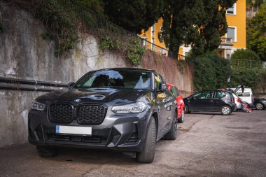 Koyu gri bir BMW SUV, sarmaşıklarla kaplı bir duvarla çevrili, yakınındaki ağaçlarla çevrili bir arazide park halinde..