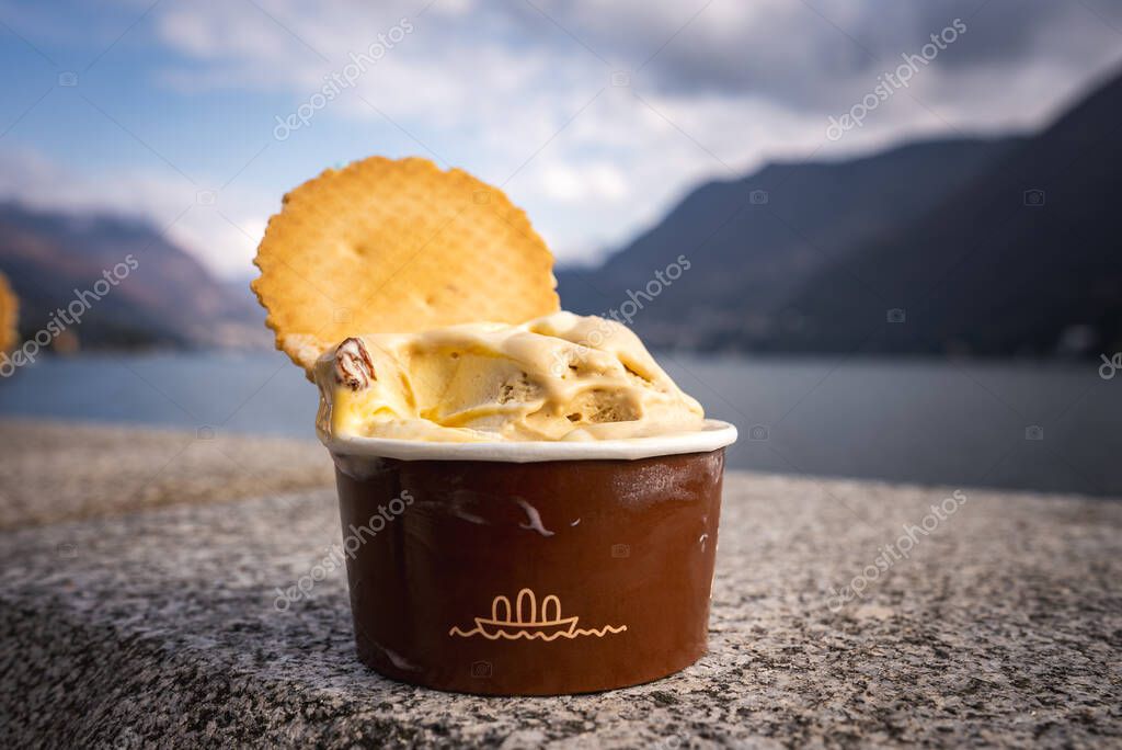A brown cup of gelato topped with a waffle biscuit sits on stone, with Lake Comos calm waters and surrounding mountains under a partly cloudy sky.