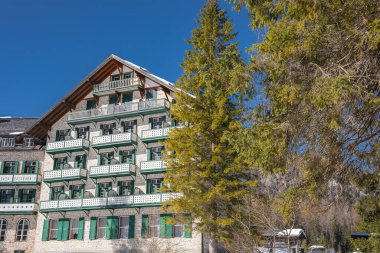 Tahta detaylar, yeşil panjurlar ve balkonlarla çevrili büyüleyici dağlık bir bina. Etrafı yemyeşil ve karla kaplı Braies Gölü, İtalya yakınlarındaki tepeler..