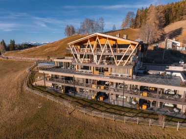 Çağdaş bir otel, üç köşeli ahşap çatılı ve cam pencereli, uzaktaki ağaçlar ve karlı dağlarla kaplı çimenli tepelere kurulmuş..