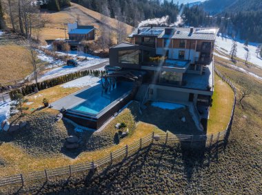 Camdan pencereleri, düz çatısı ve karlı bir dağda sonsuz havuzu olan çağdaş bir otel. Tepeler, seyrek ağaçlar ve ahşap çitlerle çevrili..