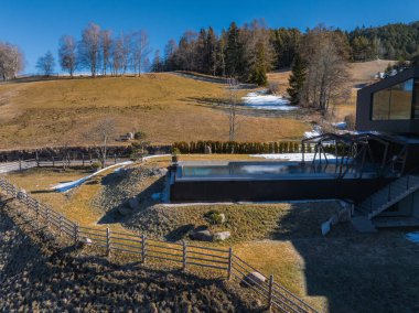 Pürüzsüz bir sonsuzluk havuzu ve engebeli tepelerin, seyrek kar parçalarının ve açık mavi gökyüzünün altında yoğun bir ormanın ortasında çağdaş bir otel mimarisi..