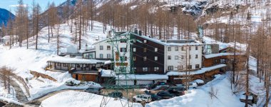Sıcak alp oteli, beyaz ve ahşap dış görünüşlü, kayak asansörü, park edilmiş arabalar, yapraksız ağaçlar, ve kışın Matterhorn yakınlarında engebeli dağlar..