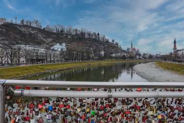 Salzburg, Avusturya, Salzach Nehri, bir aşk kilit köprüsü, Hohensalzburg Kalesi, tarihi kilise kuleleri ve parçalı bulutlu gökyüzünün altındaki çıplak ağaçlar..