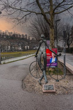 Bisiklet tekerlekleri olan ince bir insan figürü Salzburg 'da ağaçlar, tepeler ve kentsel binalarla çevrili bir nehir yolu kenarında duruyor..