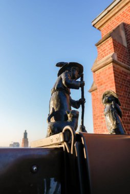 Metalik bir yüzeyde iki bronz cüce heykel, kırmızı tuğlalı bir bina ve arka planda Wroclaws eski kasabasından uzak bir kule..