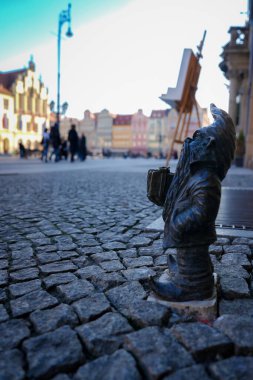 Polonya 'nın Wroclaw kentindeki kaldırım taşı caddesinde renkli tarihi binalara ve bulanık yayalara karşı küçük bir bronz cüce heykeli..