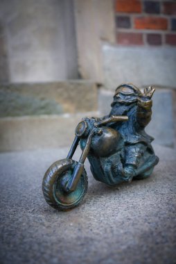 Gözlüklü, el kaldırmış, taş basamaklara dayanan ve Polonya 'nın Wroclaw şehrinde tuğla bir duvara dayanan motosikletli bir cücenin küçük bronz heykeli..