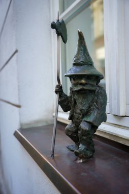 Polonya 'nın Wroclaw kentinde beyaz bir duvar ve pencereye karşı duran, sivri uçlu şapkalı ve değnekli küçük bir bronz cüce heykeli..