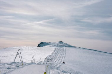 İsviçre, Zermatt 'ta görünür rayları, kayak asansörleri ve kabloları olan karlı bir kayak yamacı. Karla kaplı engebeli bir dağ zirvesi parçalı bulutlu bir gökyüzünün altında duruyor..