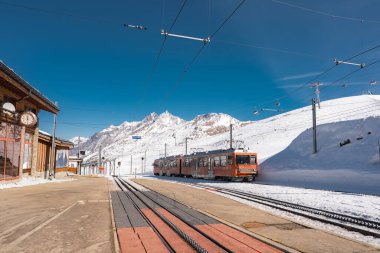 İsviçre 'nin Zermatt kentindeki karlı bir alp istasyonunda parlak turuncu bir Gornergrat tren. Matterhorn, ahşap yapılar ve açık mavi gökyüzü manzaralı..