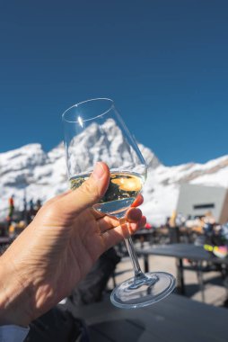 Bir el arka planda Matterhorn olan beyaz bir şarap bardağını tutuyor, karlı dağlarla çevrili, açık havada oturan, ve açık mavi gökyüzü.