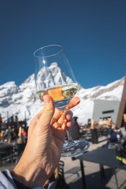 Güneşli bir günde bir el, Matterhorn 'a karşı bir bardak beyaz şarap tutuyor. Karla kaplı dağlar, masalar ve sandalyeler bir kayak merkezi ortamı yaratır..