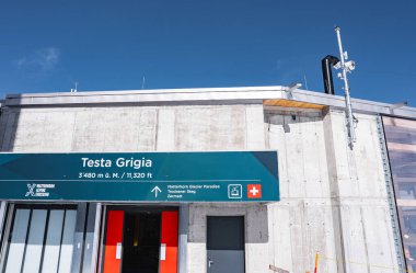 Testa Grigia istasyonunun modern beton yapısı yeşil yön işareti, İsviçre bayrağı simgesi, antenler ve yüksek irtifada açık mavi gökyüzü.