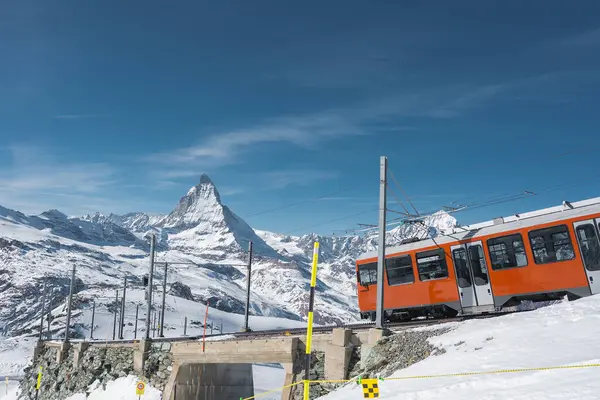 Parlak turuncu bir Gornergrat Demiryolu treni İsviçre 'nin Zermatt şehrinde kar kaplı bir taş köprüden geçer..